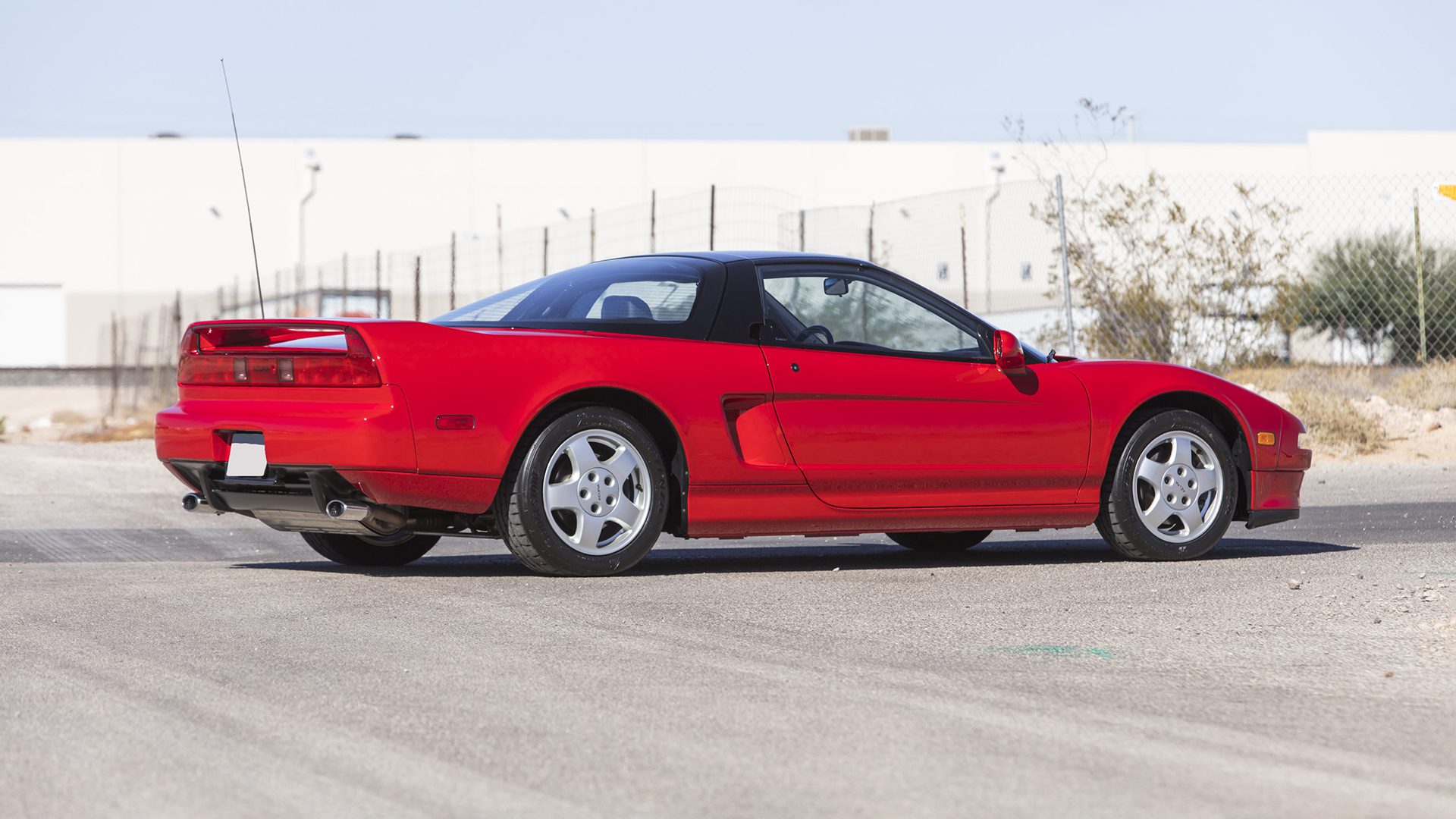 A 2022 Ferrari SF90 Spider with a striking red finish and black roof is parked on a paved surface outdoors near a fence and sparse vegetation, awaiting its moment at the Broad Arrow Las Vegas 2025 Ferrari auction.