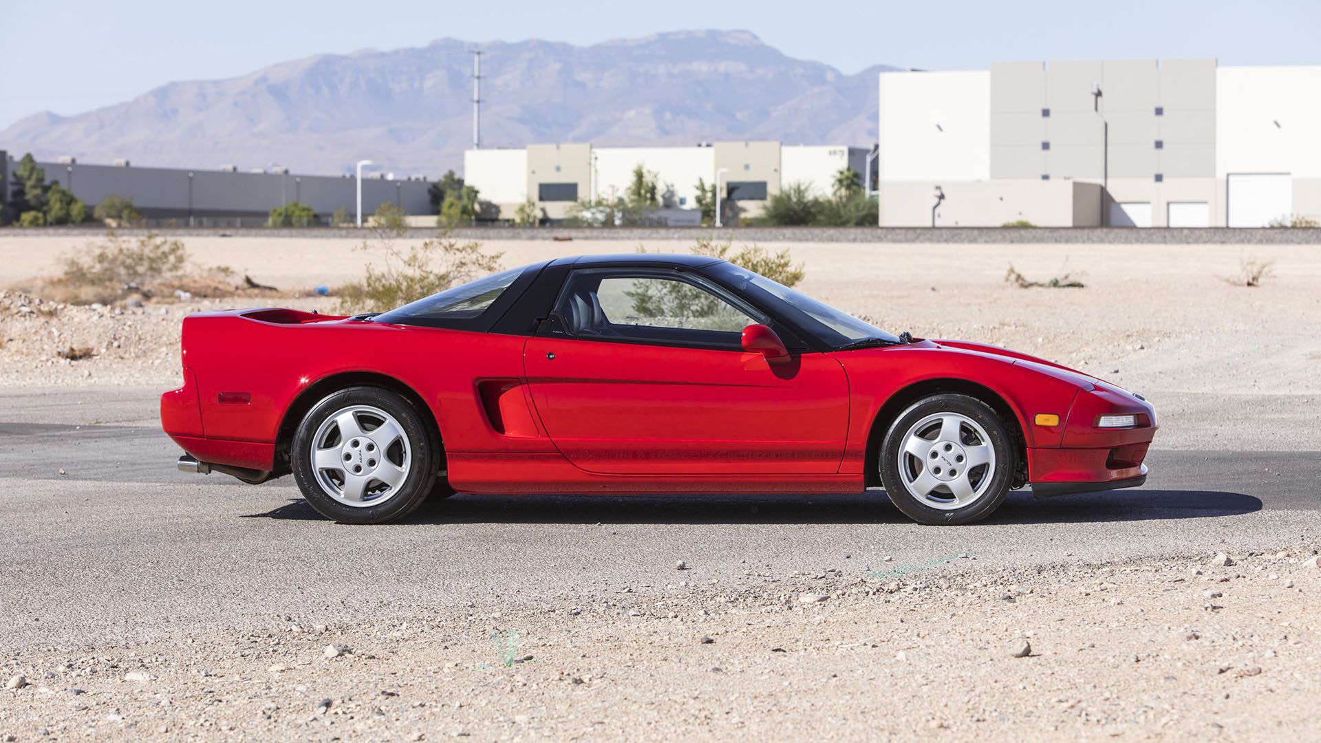 A red Ferrari SF90 Spider is parked on a paved surface in front of industrial buildings, with mountains in the background—a stunning sight ahead of the Broad Arrow Las Vegas 2025 event.