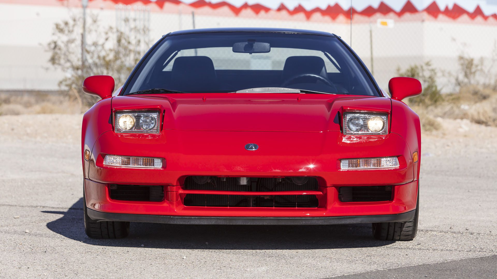 Front view of a red Acura NSX sports car with pop-up headlights, parked on a paved surface near a fence and industrial buildings—offering a 2025 Preview among icons like the Ferrari SF90 Spider at Broad Arrow Las Vegas.