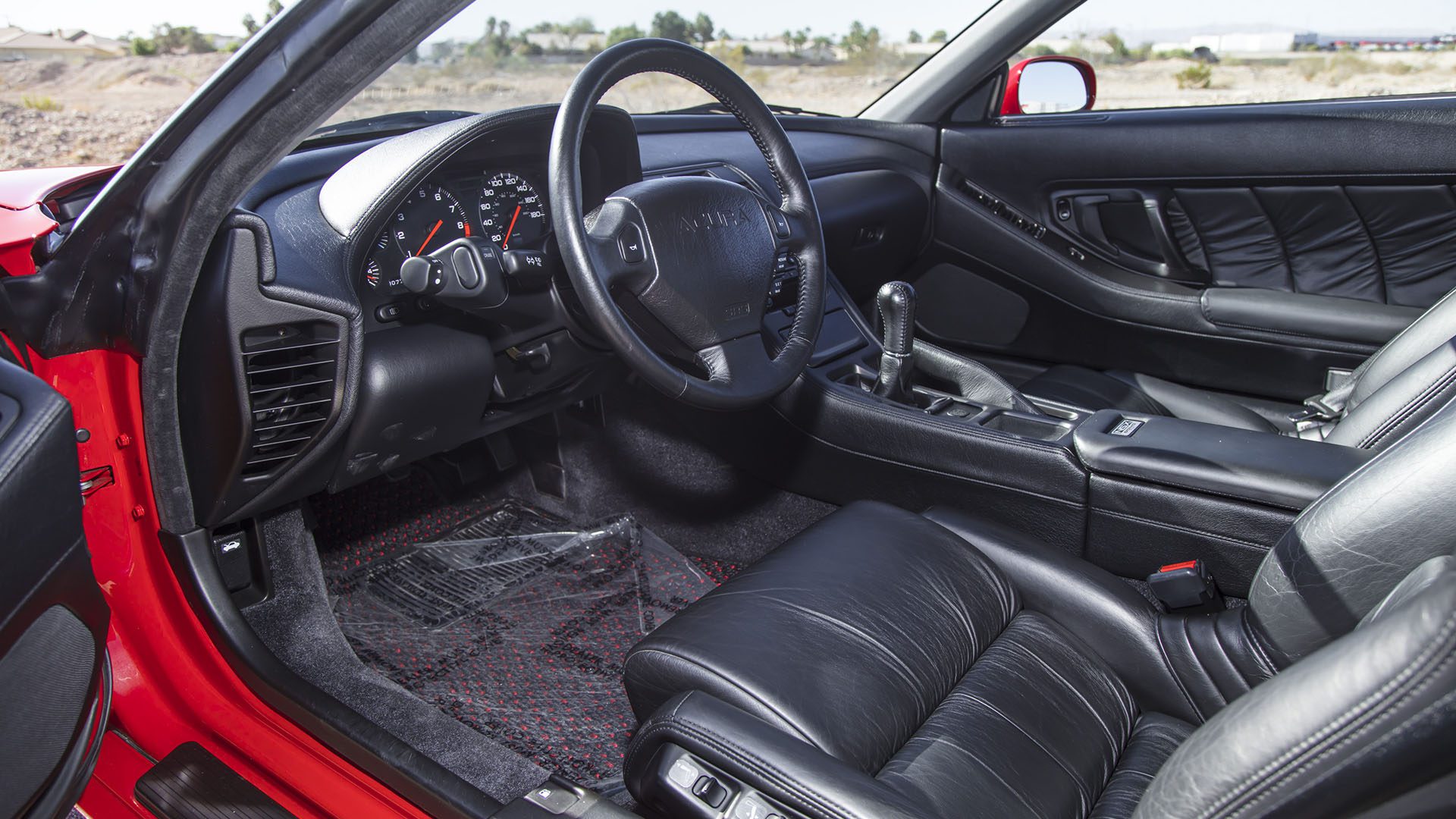 The interior of a red Ferrari SF90 Spider at the Broad Arrow Las Vegas 2025 Preview features black leather seats, a manual gear shift, and a detailed dashboard with analog gauges.