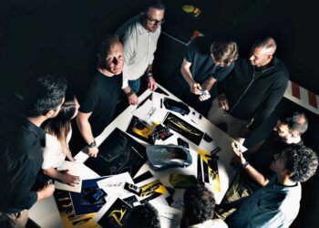 A group of people stands around a table covered with car design sketches, models, and color samples, engaged in discussion—illustrating how Centro Stile is shaping the future of Lamborghini design.