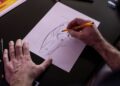 A person sketches a stylized animal head on a sheet of paper using a pencil, echoing the creative spirit of Lamborghini Centro Stile at 20: The Studio Behind Every Modern Bull, with art supplies on the black table in the background.