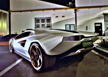 A sleek white sports car is parked at night in front of an industrial building under artificial lighting.