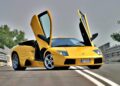 A yellow Lamborghini sports car, styled by the Lamborghini Centro Stile at 20: The Studio Behind Every Modern Bull, sits on a road with its scissor doors open against a blurred backdrop of trees and railings.