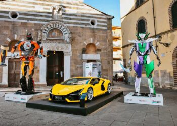 A yellow sports car is displayed between two giant robot sculptures at Lucca Comics & Games, where Lamborghini turns factory waste into impressive robots, set against the backdrop of historic buildings in an outdoor plaza.