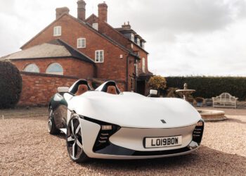 A white, two-seater sports car with license plate LO1980M is parked on a gravel driveway in front of a large brick house.