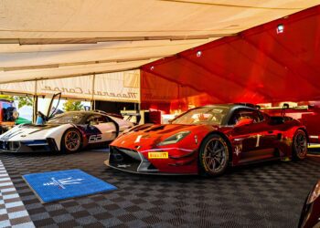 Two Maserati sports cars, one red and one white, are parked inside a tented area with checkered flooring and branded mats as the Maserati GT2 debuts stateside at GT America in Indianapolis.