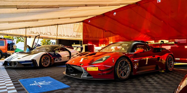 Two Maserati sports cars, one red and one white, are parked inside a tented area with checkered flooring and branded mats as the Maserati GT2 debuts stateside at GT America in Indianapolis.