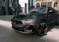 A dark gray Maserati Grecale Folgore SUV, featuring advanced EV tech, is parked on a concrete surface in front of modern glass buildings and a historic stone building.