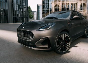 A dark gray Maserati Grecale Folgore SUV, featuring advanced EV tech, is parked on a concrete surface in front of modern glass buildings and a historic stone building.