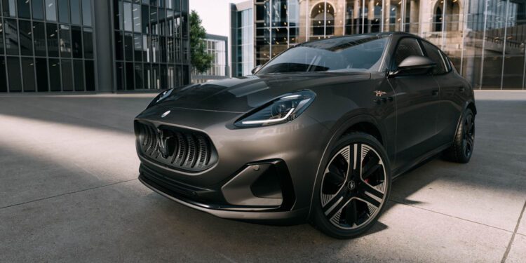 A dark gray Maserati Grecale Folgore SUV, featuring advanced EV tech, is parked on a concrete surface in front of modern glass buildings and a historic stone building.