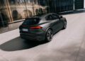 A matte gray Maserati Grecale Folgore SUV, featuring advanced EV tech, is parked on a modern, sunlit plaza beside a glass building reflecting nearby architecture.