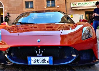 A red Maserati sports car, showcasing MCPura key design elements, is parked on a cobblestone street, with a man nearby using his phone and a building in the background. Q&A: Maserati’s Mastermind Reveals These Standout Features.