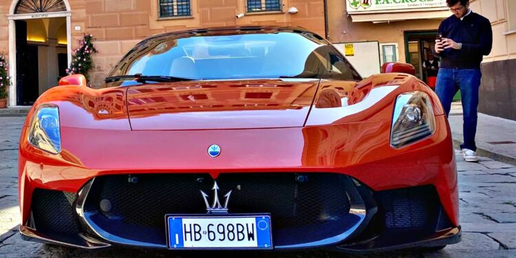 A red Maserati sports car, showcasing MCPura key design elements, is parked on a cobblestone street, with a man nearby using his phone and a building in the background. Q&A: Maserati’s Mastermind Reveals These Standout Features.