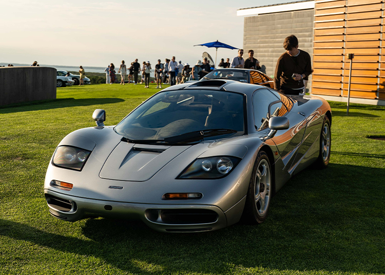 A silver sports car, an icon of automotive art, is parked on grass with people gathered in the background on a sunny day.