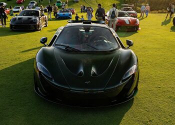 A black McLaren sports car, a true piece of automotive art, is displayed on a grassy field among other luxury vehicles and guests at the exclusive Bridge IX outdoor car event.