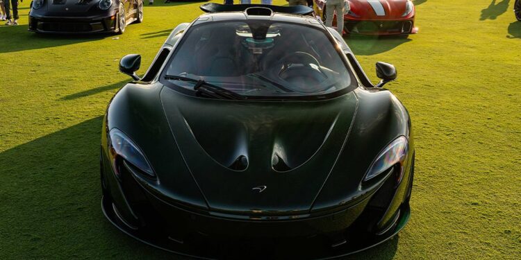 A black McLaren sports car, a true piece of automotive art, is displayed on a grassy field among other luxury vehicles and guests at the exclusive Bridge IX outdoor car event.