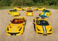 Eight yellow Ferrari sports cars with white and blue racing stripes, part of exclusive Ferrari Collections, are arranged in a semicircle on a gravel surface, with trees and grass in the background.