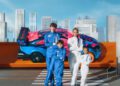 Four people in blue and white racing suits stand on a crosswalk in front of a Mercedes-Benz CLA that gets a wild, life-size Hot Wheels makeover, with city buildings adding to the vibrant scene.