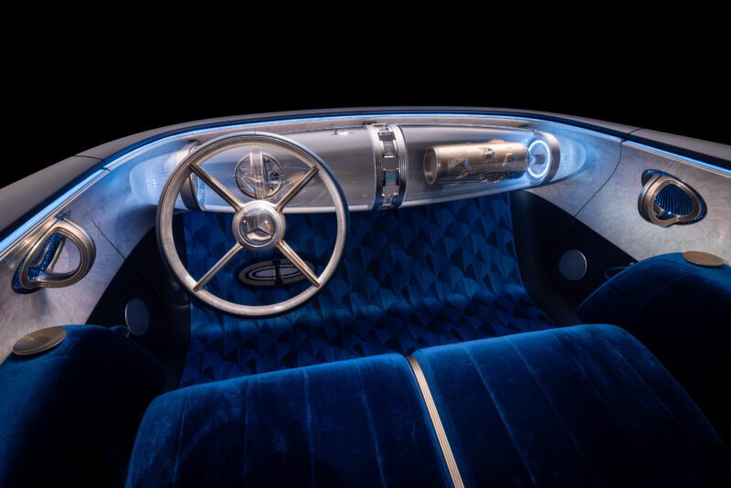 A futuristic car interior inspired by the Mercedes-Benz Vision Iconic revives classic elegance for the digital era, with a metallic steering wheel, blue geometric-patterned seats, and a sleek, illuminated dashboard.