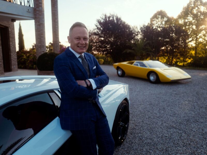 A man in a blue suit stands with arms crossed, leaning against a white car, with a yellow Lamborghini Centro Stile at 20: The Studio Behind Every Modern Bull sports car parked in the background on a gravel driveway.