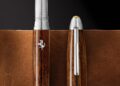 Two wooden pens with silver accents and a prancing horse logo, inspired by Ferrari, stand upright on brown leather, with one pen's cap removed and placed beside it.