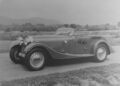 Black and white photo of a vintage convertible car parked on a rural road with fields and hills in the background.