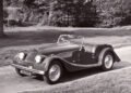 A classic Morgan convertible car is parked on a road with trees and grass in the background.
