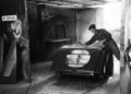 A worker in overalls and a cap spray paints a Morgan Plus Four car body panel in an industrial workshop with a "No Smoking" sign on the wall, celebrating Morgan’s 75th Anniversary.