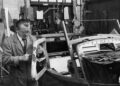 A man in a workshop uses a tool to measure a wooden car frame for a Morgan Plus Four next to a partially assembled vintage vehicle, reflecting Morgan's craftsmanship in celebration of its 75th Anniversary.
