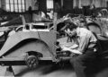 A man assembles the frame of a Morgan Plus Four in an industrial workshop, surrounded by car parts and machinery, celebrating the 75th Anniversary of this iconic vintage model.