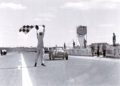 A race official waves a checkered flag as a vintage Morgan Plus Four crosses the finish line on a track, celebrating the 75th Anniversary amid cheering spectators and colorful advertisements in the background.