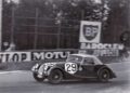 Black-and-white photo of a vintage Morgan Plus Four race car labeled number 29 driving on a track, with advertising signs and trees in the background—celebrating Morgan's 75th Anniversary.