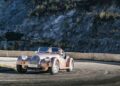 A classic Morgan Plus Four anniversary convertible with two people inside drives on a winding mountain road, rocky slopes and sparse trees lining the background, celebrating 75th Anniversary automotive heritage.