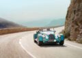 A classic-style Morgan Plus Four blue convertible sports car drives on a winding mountain road with rocky cliffs and hazy hills in the background.