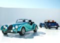 Two vintage-style convertible cars, including a Morgan Plus Four 75th Anniversary edition, are parked on a glossy white surface—one facing forward, the other away—set against a minimal, sunlit backdrop.