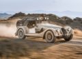 A vintage-style Morgan Plus Four rally car drives at speed on a dusty dirt road, kicking up dust with blurred hills and trees in the background, celebrating Morgan’s 75th Anniversary spirit.