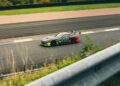 A camouflaged race car with neon yellow and red accents drives on a racetrack, viewed from behind a guardrail.