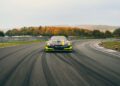 A camouflaged Mercedes AMG TRACK SPORT concept sports car drives on a wet racetrack surrounded by autumn trees under a cloudy sky.