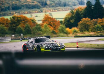 A camouflaged Mercedes CONCEPT AMG TRACK SPORT with yellow accents speeds along a racetrack, weaving through circuits bordered by trees in vibrant autumn foliage.