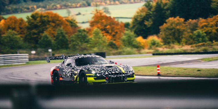 A camouflaged Mercedes CONCEPT AMG TRACK SPORT with yellow accents speeds along a racetrack, weaving through circuits bordered by trees in vibrant autumn foliage.