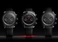 Three black Omega Speedmaster chronograph watches are displayed side by side against a dark background, with the center watch featuring a red second hand.