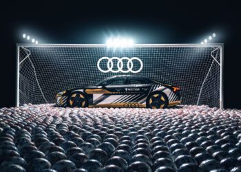A black and gold Audi sports car is parked in front of a goalpost filled with soccer balls under bright stadium lights, celebrating the Audi partnership with Major League Soccer, with an illuminated Audi logo above the net.