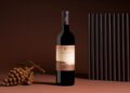 A bottle of Il Bruciato red wine stands upright beside a bunch of dried grapes and a vertical black ridged panel, set against a brown background—a refined scene reminiscent of Maserati’s Grecale Tributo elegance.