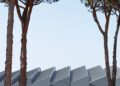 Two tall pine trees frame a modern building with angular, zigzag-shaped roofs and grey walls—its sleek lines echoing the style of a Maserati—set against a clear sky and surrounded by greenery.