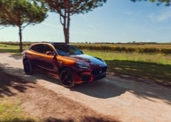 A Maserati Grecale Tributo red SUV drives on a dirt road lined with trees under a clear blue sky, with fields visible in the background.