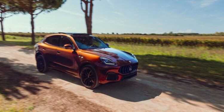 A Maserati Grecale Tributo red SUV drives on a dirt road lined with trees under a clear blue sky, with fields visible in the background.