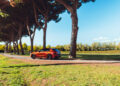 A red Maserati Grecale Tributo SUV drives on a tree-lined path in a sunny, green park with clear blue skies.