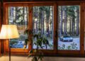 View through a window showing a parked Maserati sports car among tall trees, with a lit lamp and a potted plant indoors in the foreground—capturing an elegant Italian ambiance.