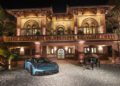 A blue Maserati sports car and a black grand piano are displayed in front of a large, ornate brick mansion in Italy, illuminated at night, evoking the elegance of the MCPURA Driving Experience.